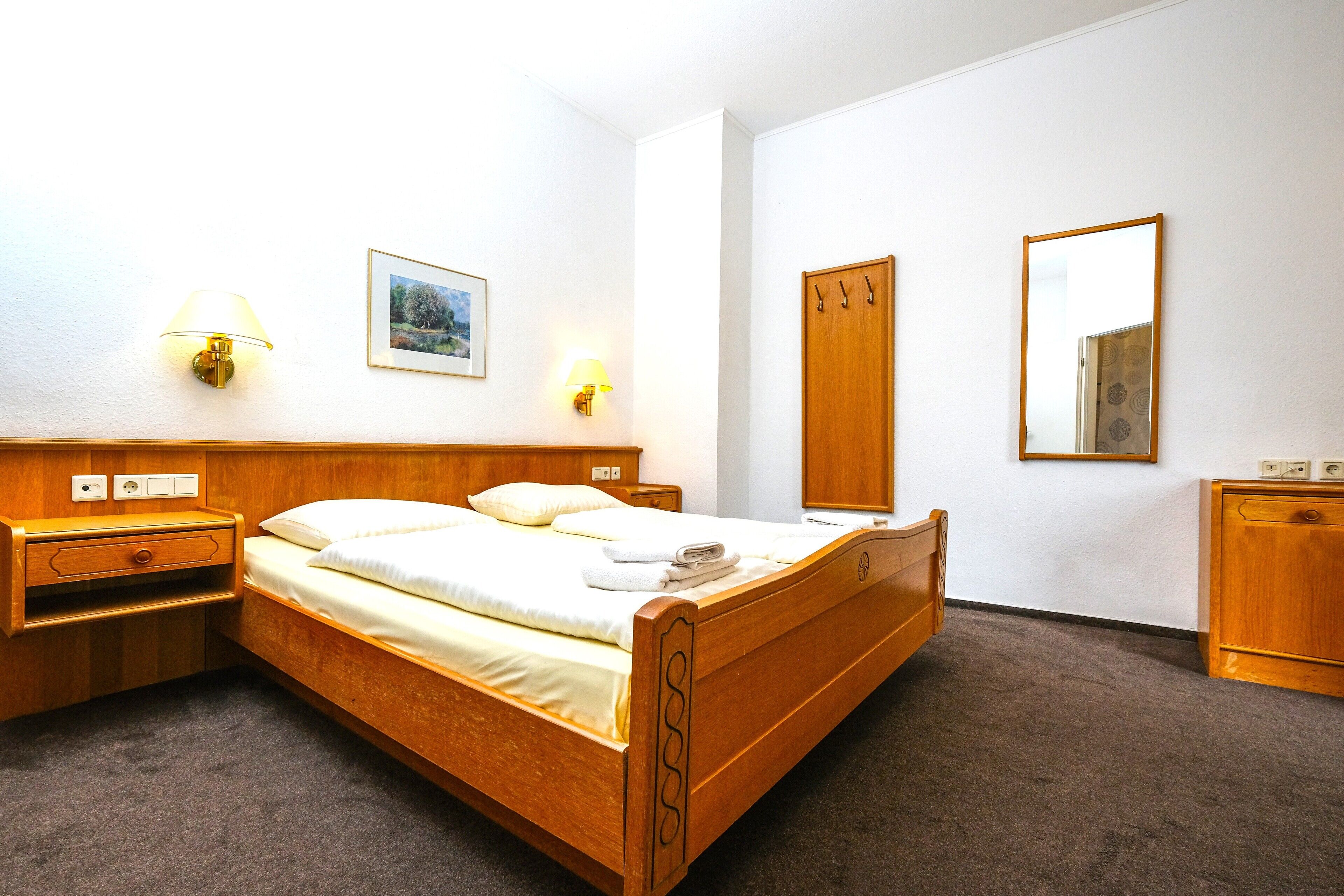 A wooden bed with a headboard, bedside tables, a nightstand, a lamp, a framed picture, a mirror, and a wardrobe.