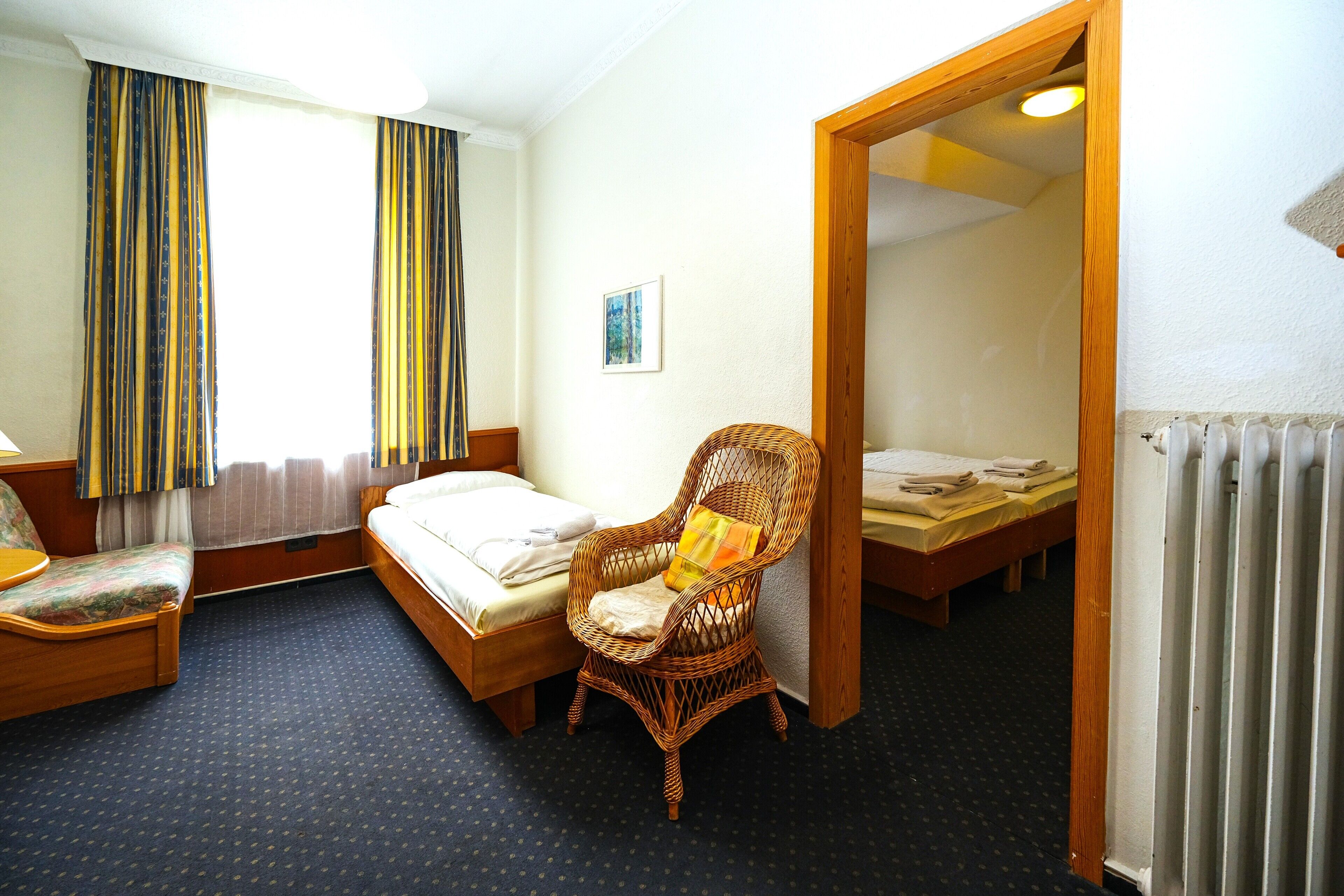 Quarto de hotel com uma cama de solteiro, uma cadeira, uma escrivaninha e uma janela grande com cortinas.