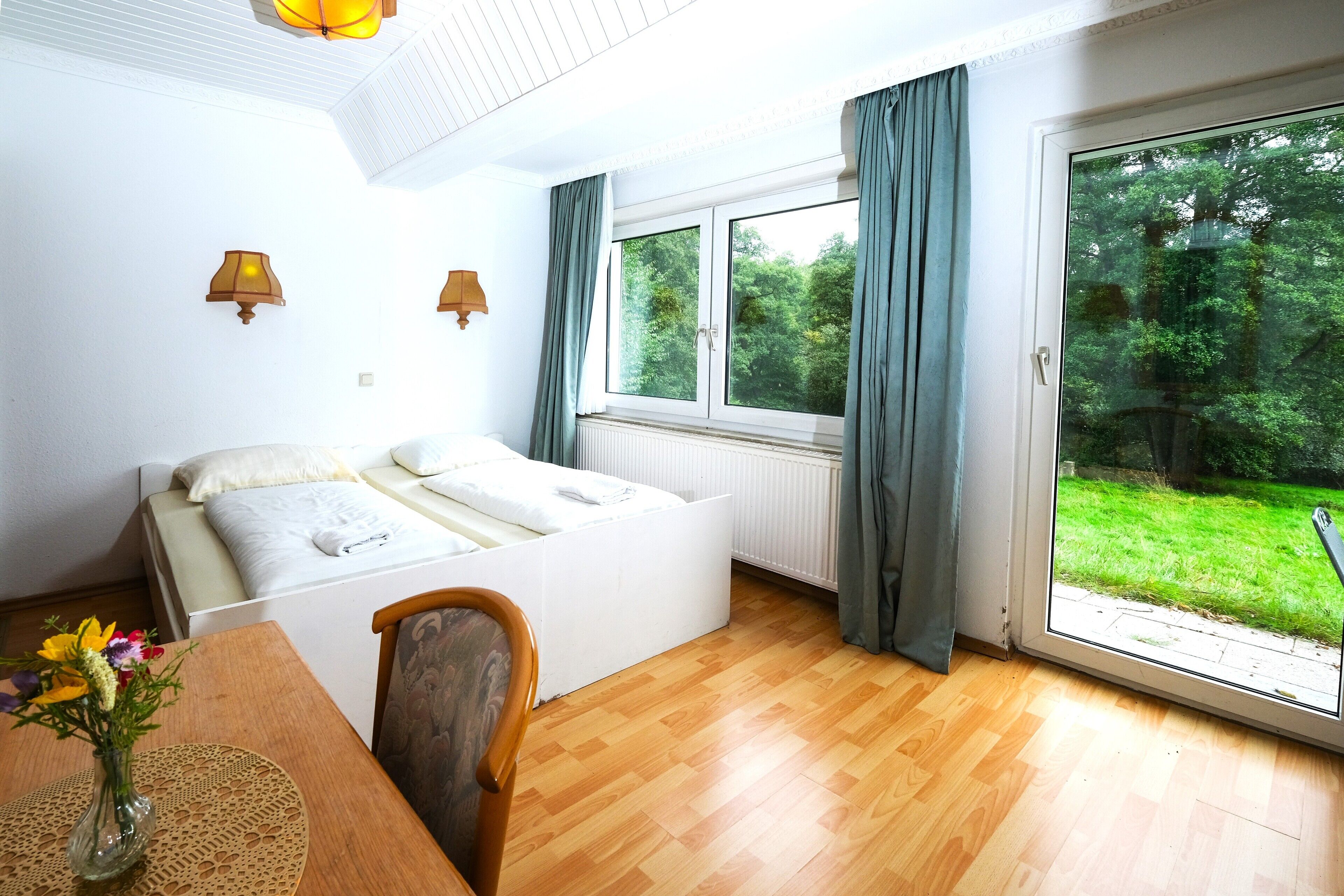 Ein Schlafzimmer mit Bett, Fenster, Blick ins Grüne und einem Tisch mit Blumen.