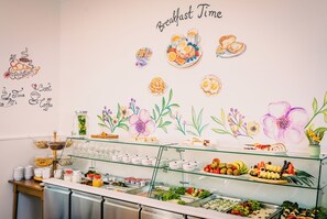 Un buffet de petit-déjeuner proposant une variété d’aliments, et un mur décoré d’illustrations florales ainsi que de l’inscription « Heure du petit-déjeuner ».