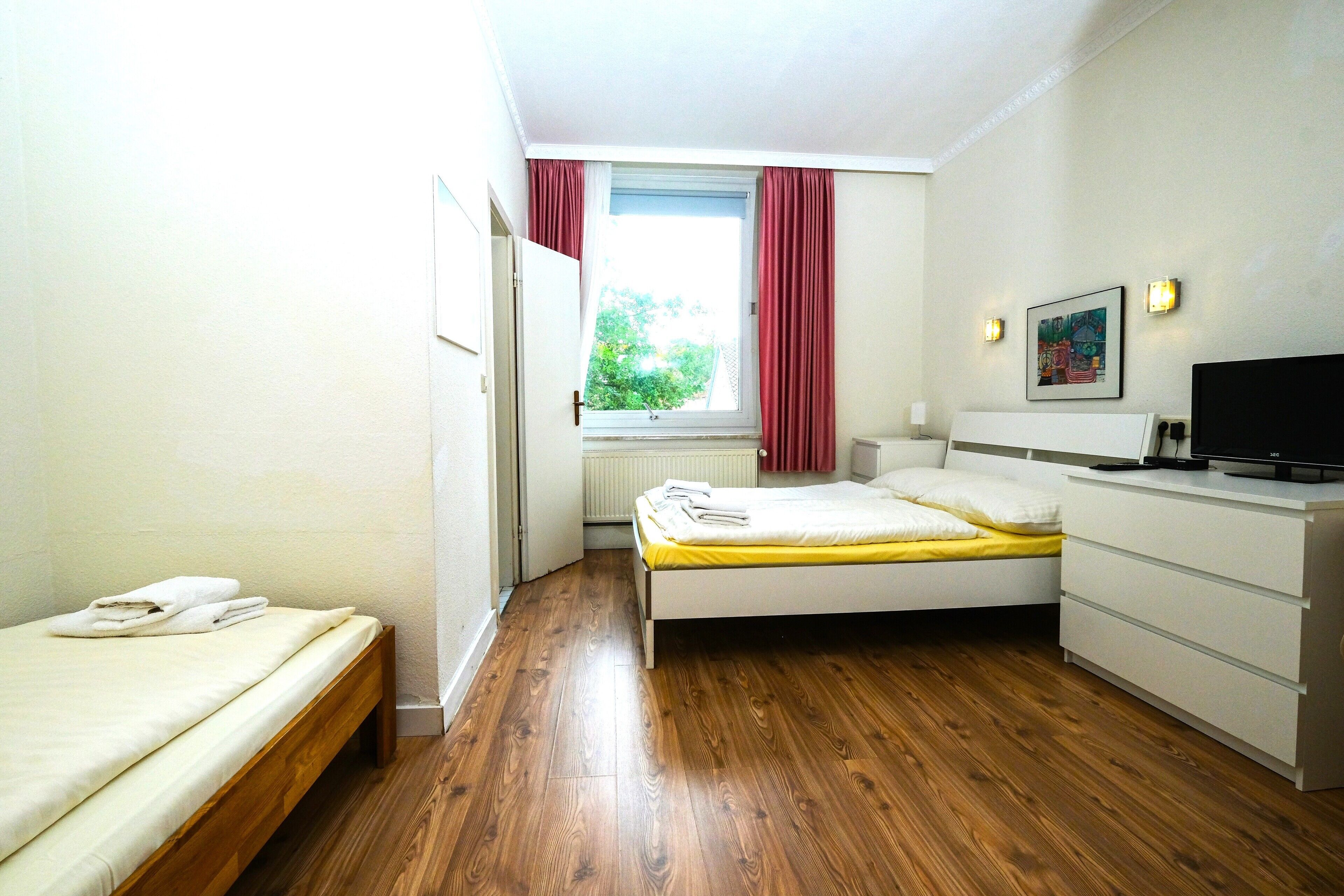 Quarto pequeno e bem iluminado com piso de madeira, uma cama de solteiro com colchão amarelo, mesa de cabeceira branca, televisão e janela com cortinas vermelhas.