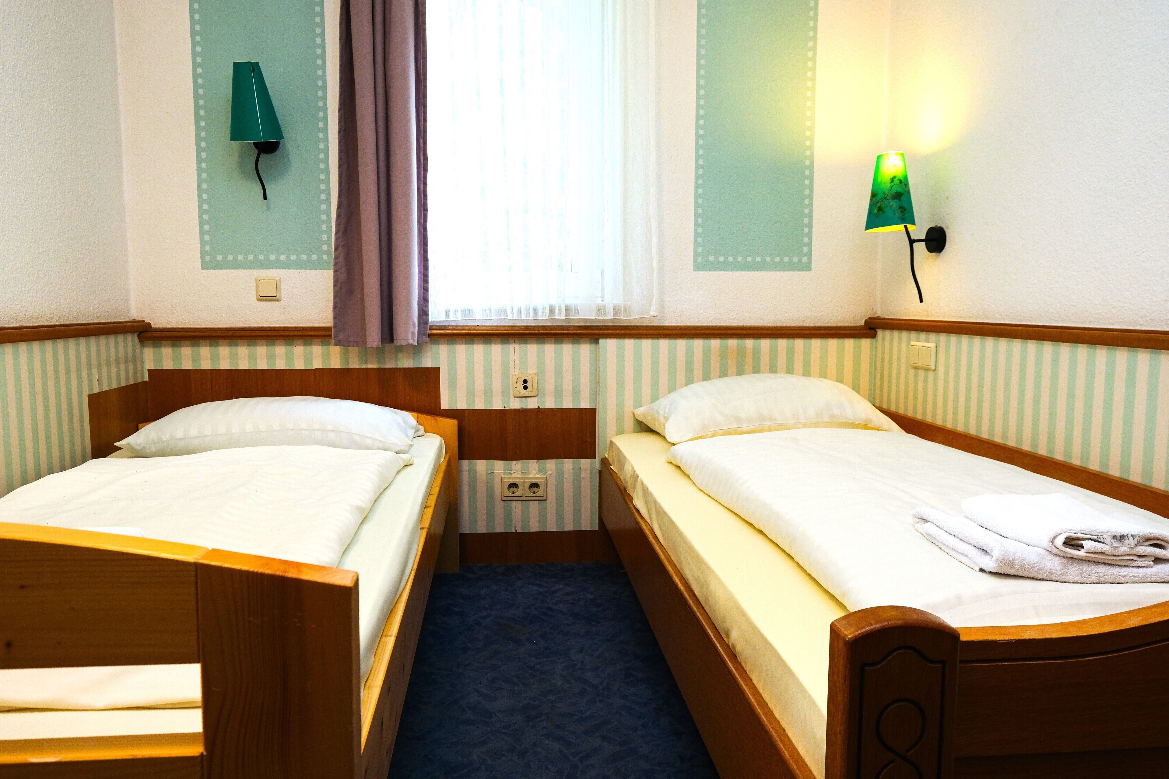 Een kamer met twee houten bedden, een raam met gordijnen en een wandlamp.