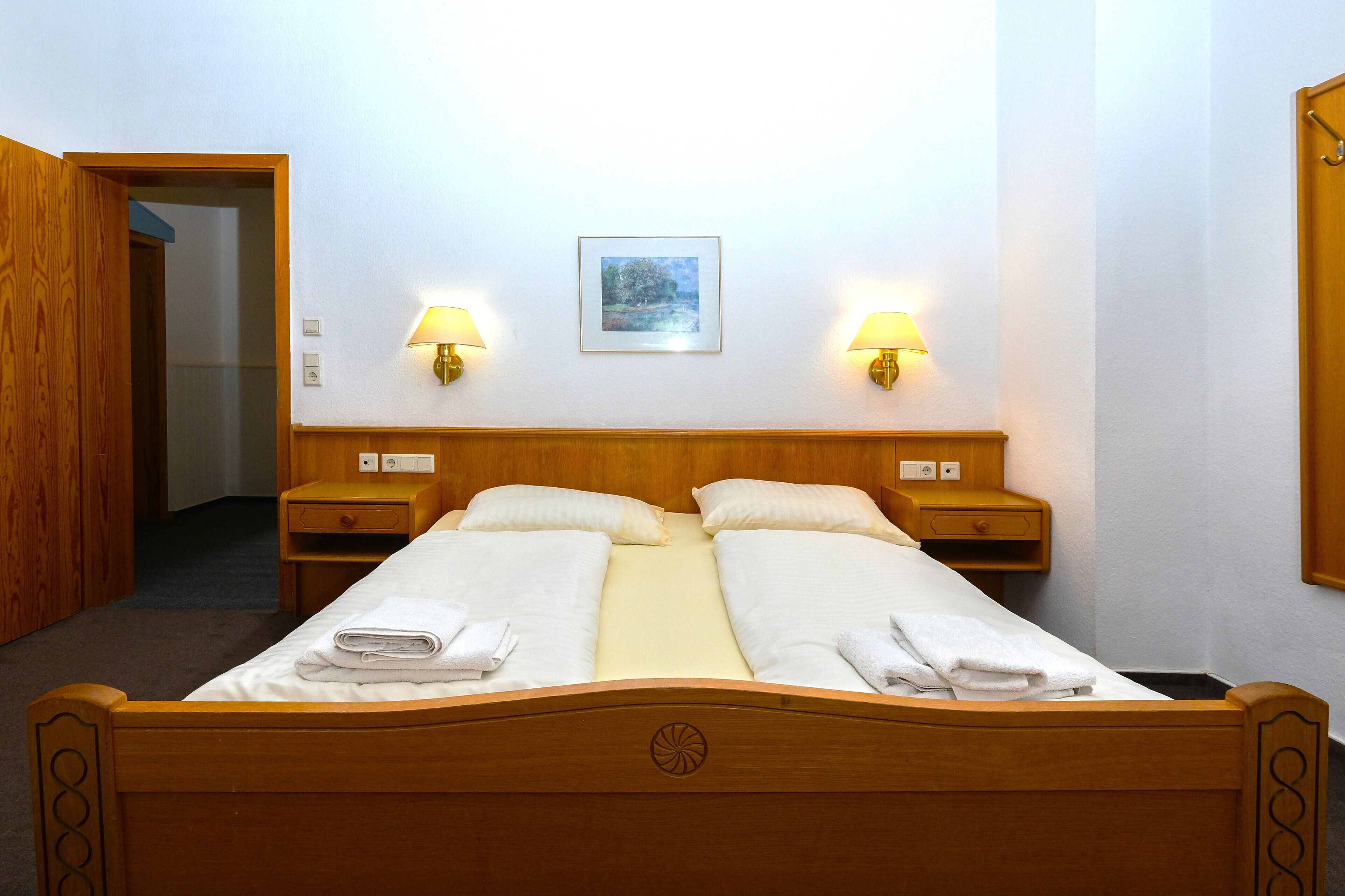 A neatly made double bed with white linens, two bedside tables with lamps, a framed picture on the wall, and a wooden door leading to another room.