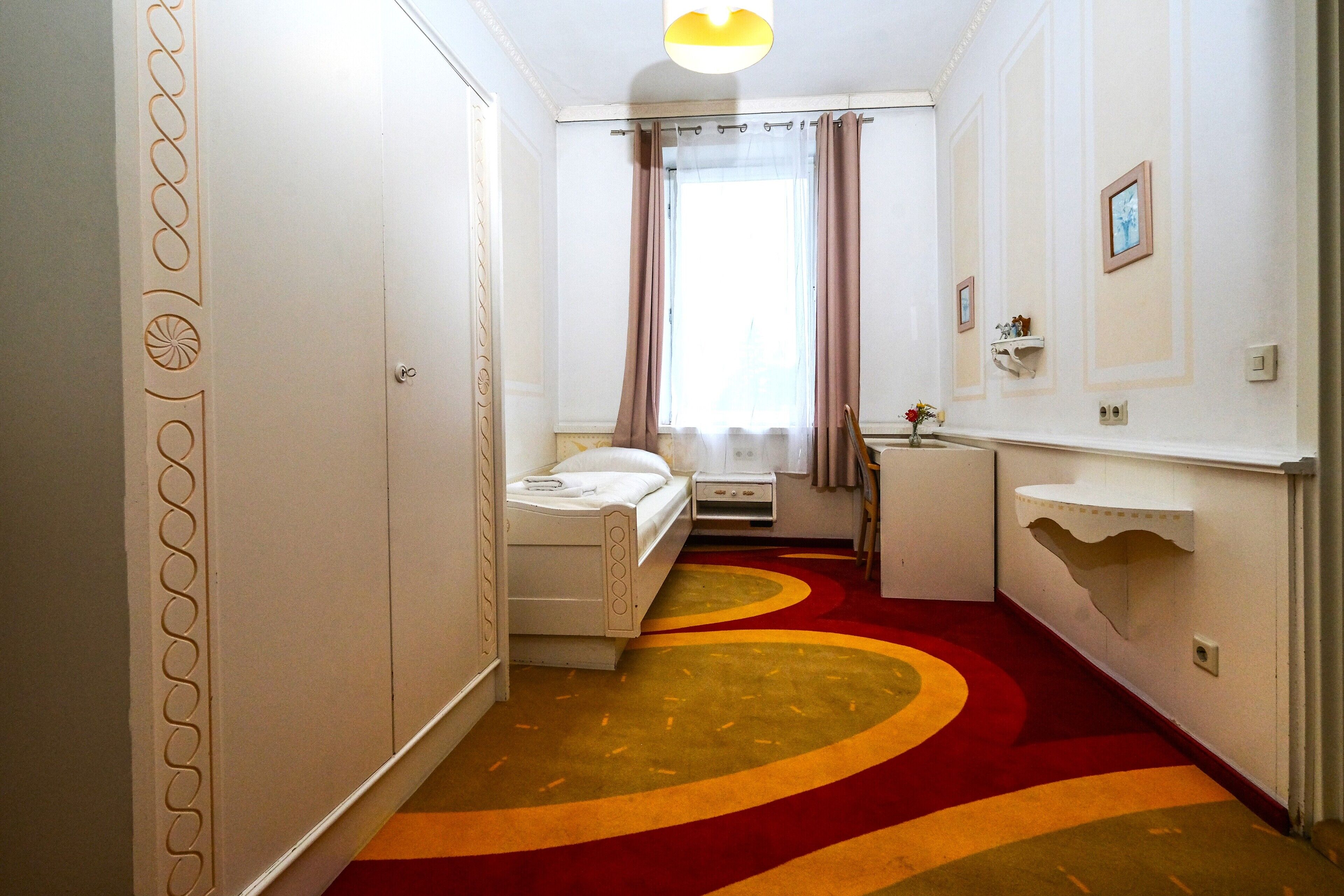 Habitación de hotel con cama, mesita de noche, escritorio pequeño, silla, ventana con cortinas, estante en la pared y alfombra decorativa.