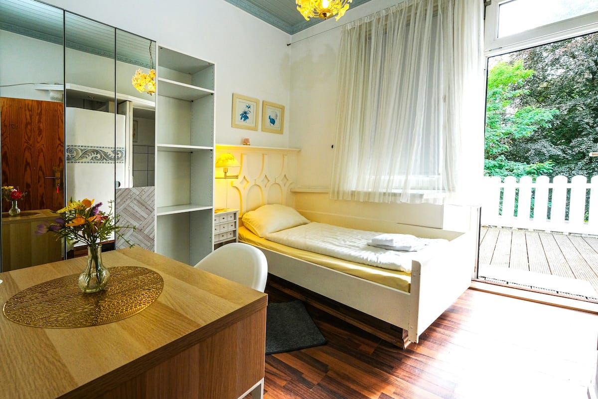 Quarto com cama, escrivaninha com um vaso de flores, cadeira e janela com vista para vegetação.