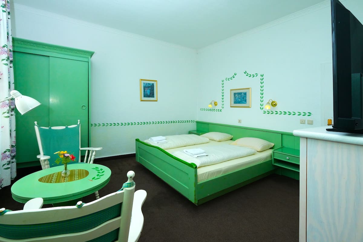 Quarto com cama verde, uma pequena mesa redonda com abajur, uma cadeira e uma televisão sobre um suporte.