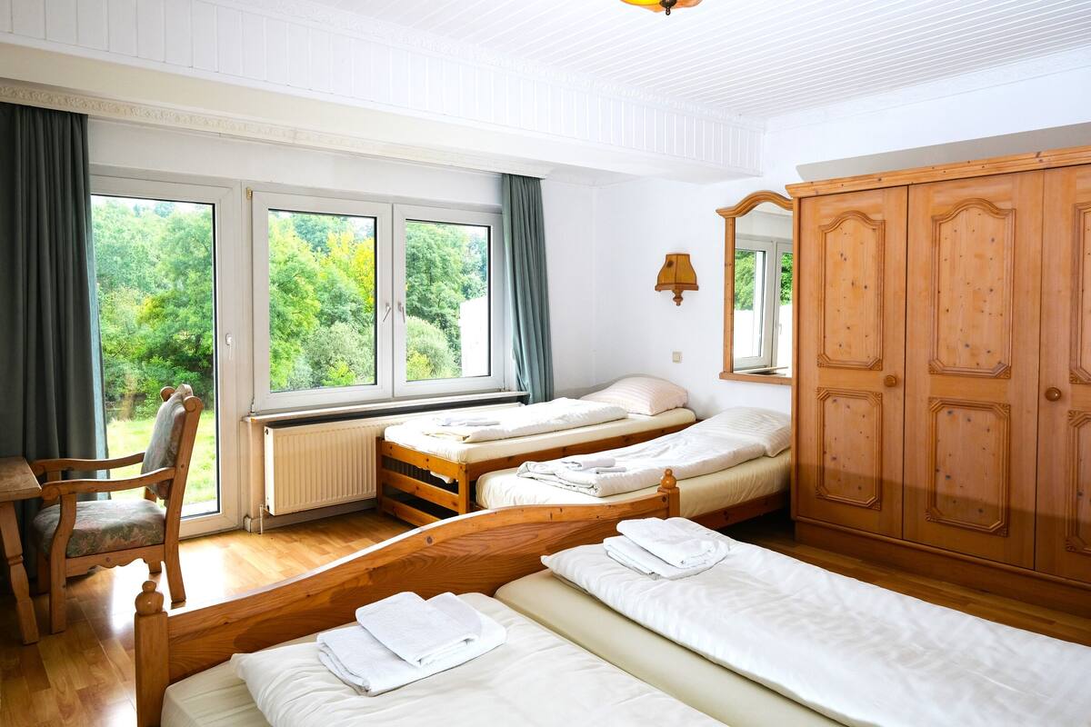 Quarto com três camas de madeira, um guarda-roupa de madeira, uma cadeira e uma janela com cortinas.