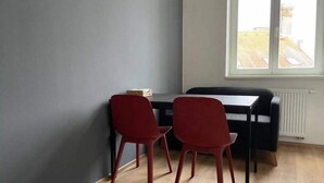A room with a wooden floor, a grey wall, and a window with a view of buildings. There is a black table with two red chairs and a small box on it. - Brand New Apartments 4.1 (Hlavní mesto Praha)