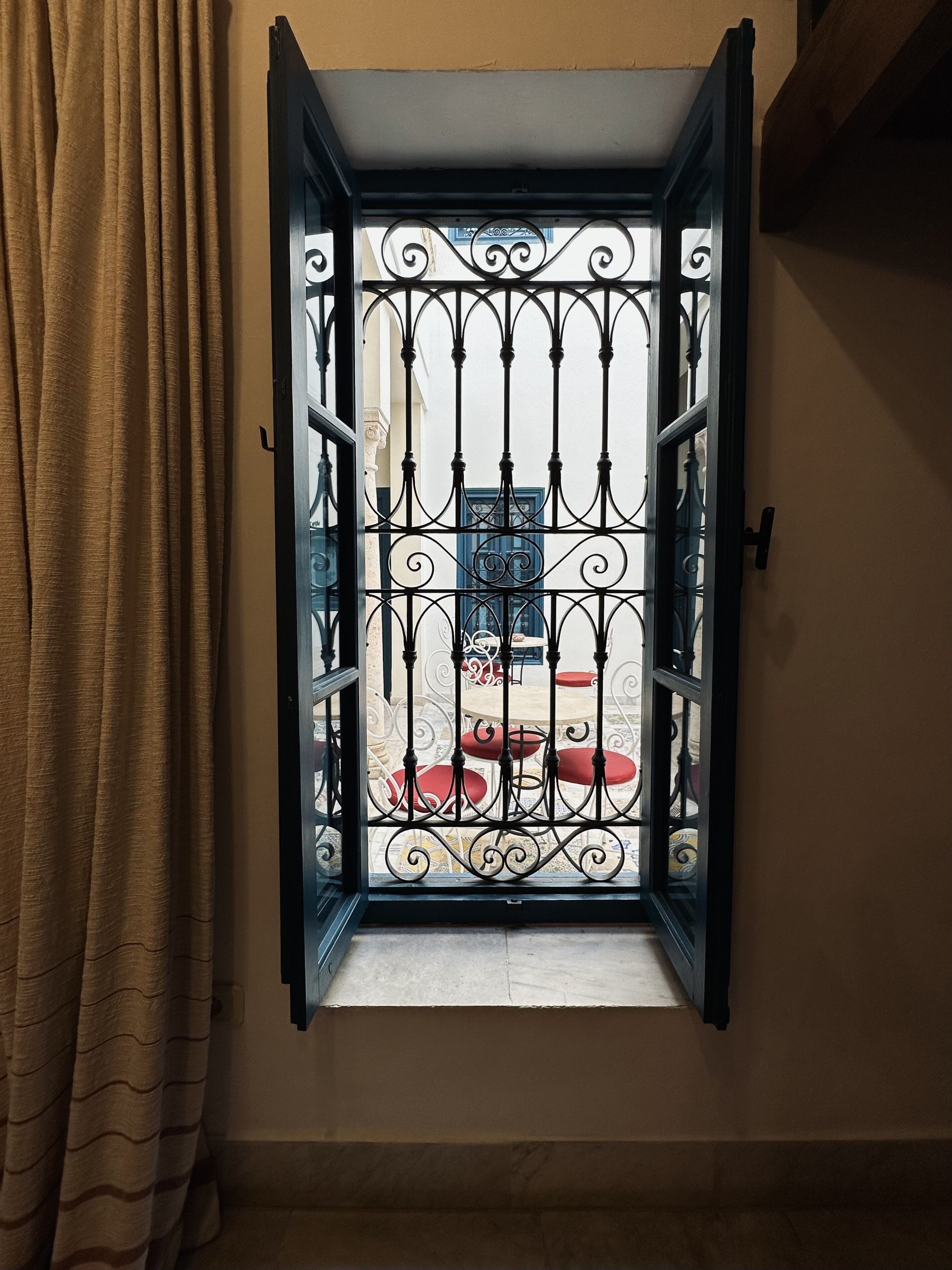 A window with ornate black metal bars, a view of a balcony with red cushions, and a beige curtain partially drawn to the side.