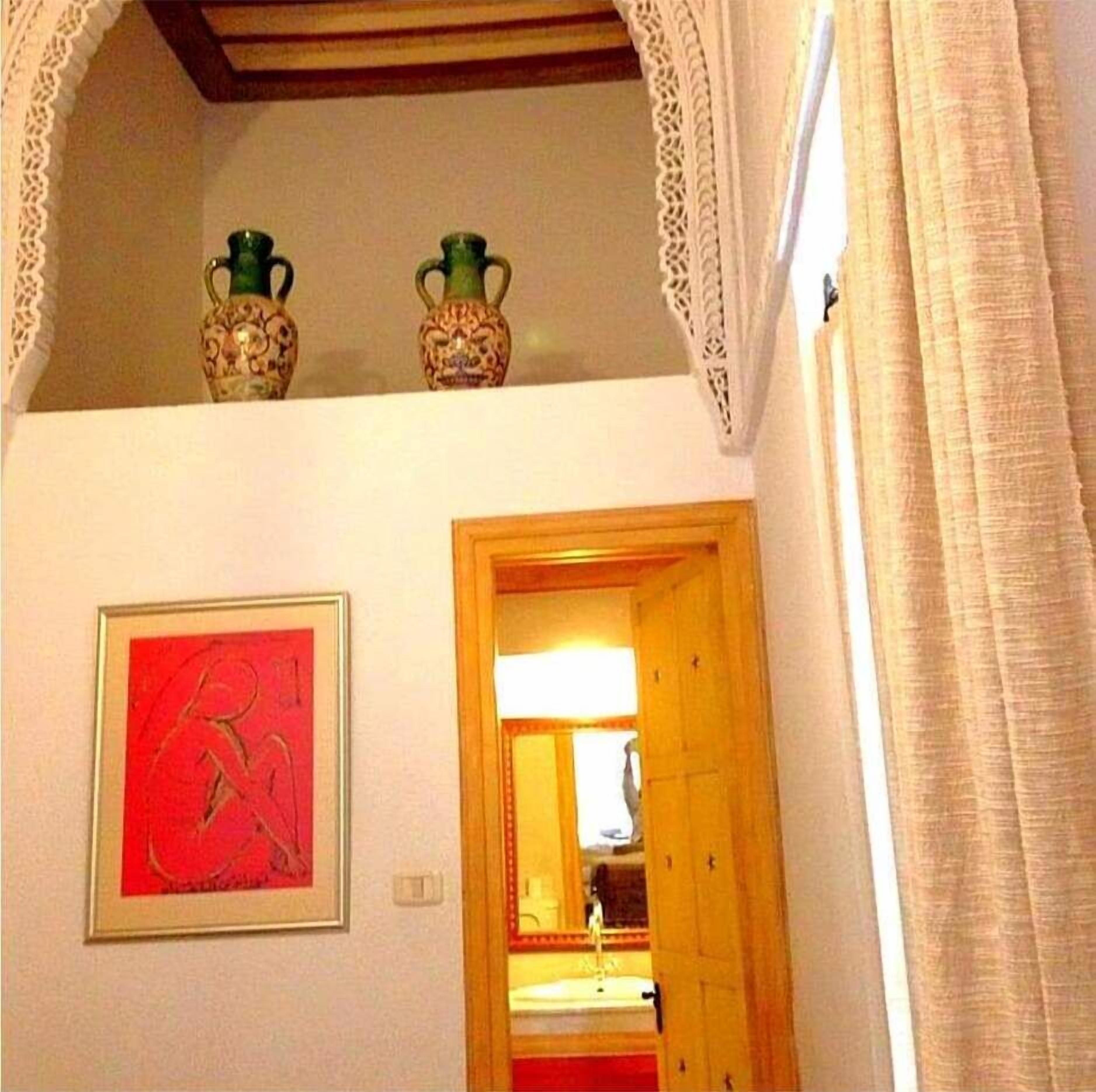 Una habitación con un cuadro abstracto enmarcados, una puerta de madera que da a otra estancia, y dos jarrones decorativos en el techo.