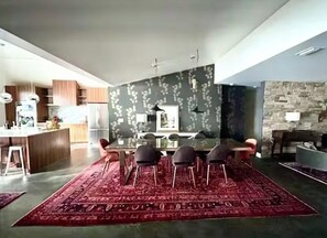 A spacious living area with a dining table and chairs, a kitchen in the background, and a patterned rug on the floor.