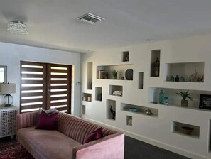 Uma sala de estar com um sofá, uma estante fixada na parede com objetos decorativos e uma porta com ripas horizontais.