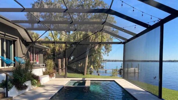 A swimming pool with a glass enclosure, surrounded by a deck and a view of a lake.