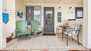 Un porche con dos sillas Adirondack verdes, una pequeña mesa redonda y un banco. Hay una puerta con un ventanal encima, y un letrero que dice "Welcome to the Back Porch".