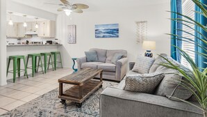 Una sala moderna con un sofá gris, una mesa de centro de madera y una cocina con armarios blancos y taburetes verdes.
