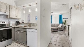 Cocina moderna con encimeras de granito, electrodomésticos de acero inoxidable e iluminación colgante. Se aprecia, al fondo, una zona de comedor con sofá y mesa de centro.