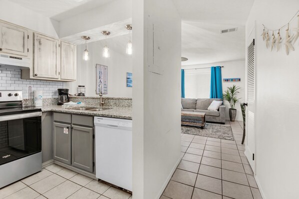 A modern kitchen with granite countertops, stainless steel appliances, and pendant lighting. A dining area with a sofa and coffee table is visible in the background.