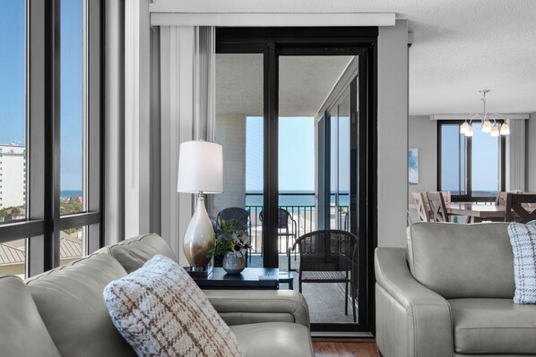A modern living room with a sofa, a table with a lamp, and a view of the balcony and ocean.