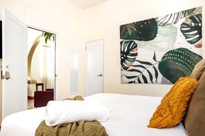 Een hotelkamer met een bed, een schilderij van tropische bladeren en uitzicht op een badkamer met een spiegel en een rode stoel.