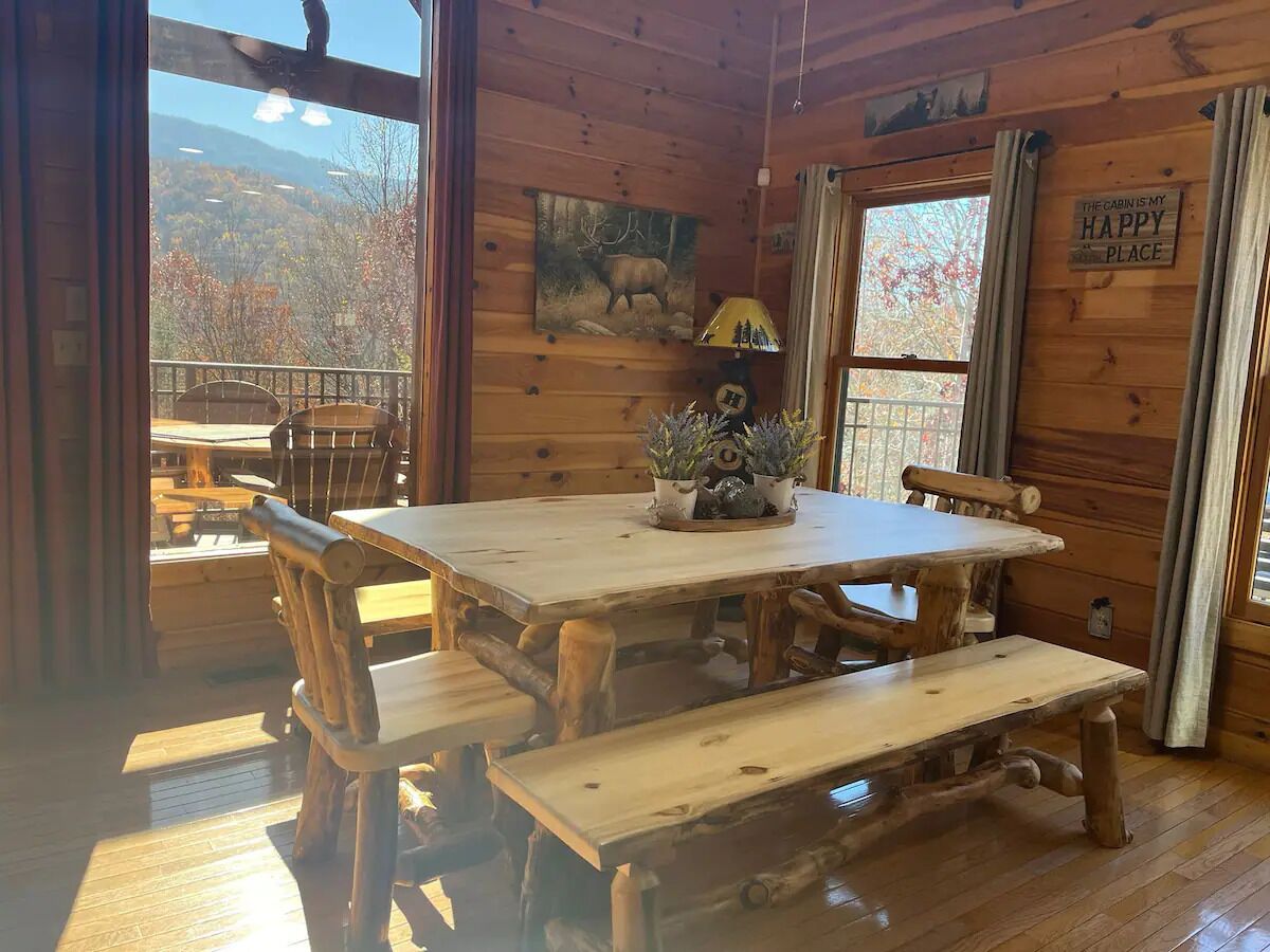 A rustic cabin interior with a wooden dining table, benches, and a view of the outdoors.