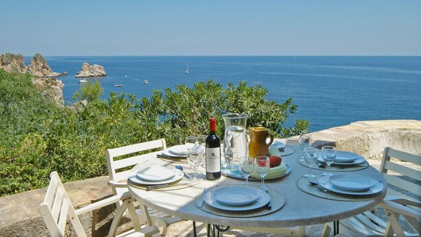 A table set for four with a bottle of wine, glasses, and a pitcher, overlooking a coastal view with boats and rocky terrain. - Villa La Fenice by Wonderful Italy (Castellammare del Golfo)