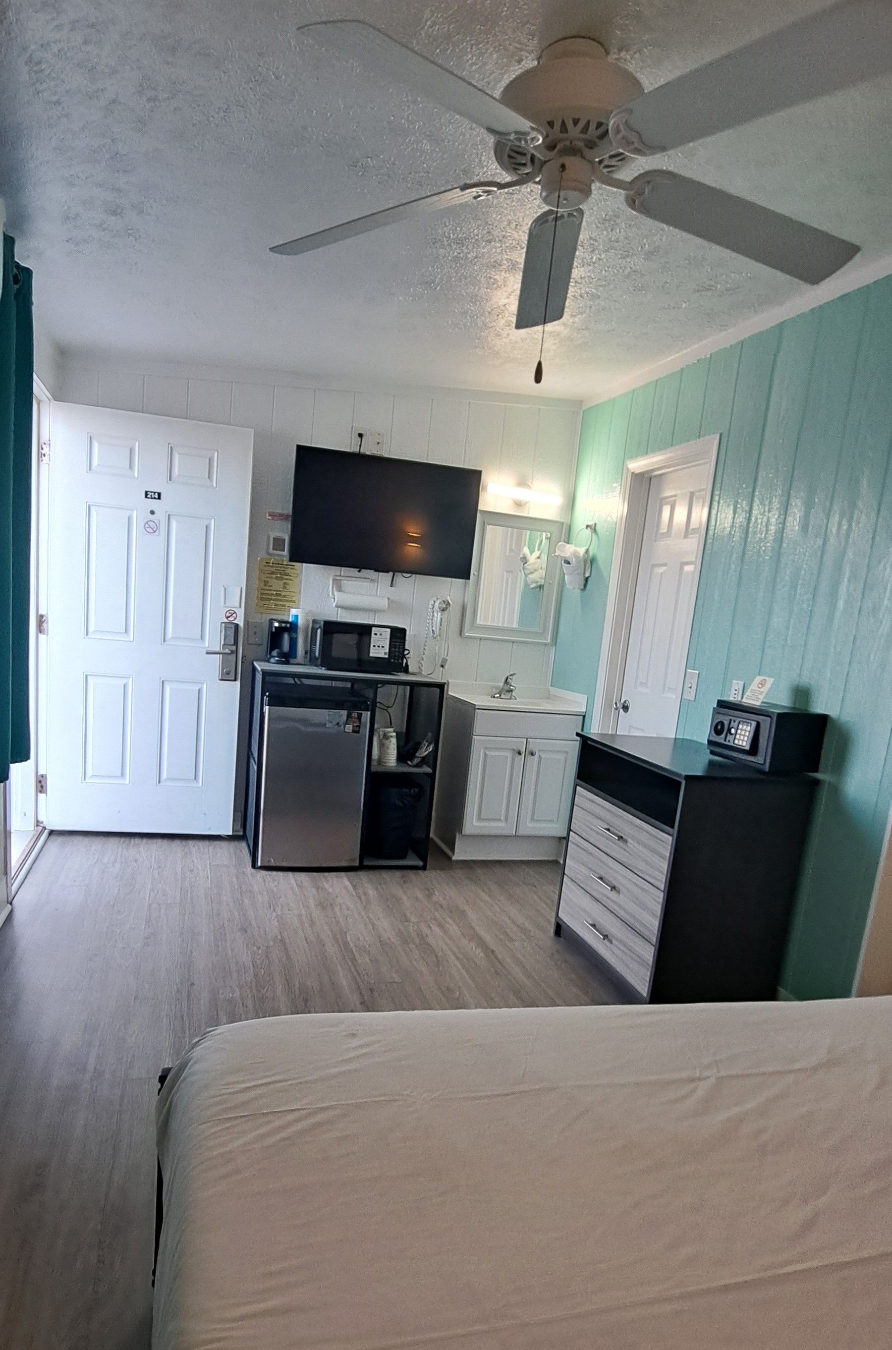 Una habitación de hotel compacta con kitchenette, una cama, ventilador de techo y televisor.