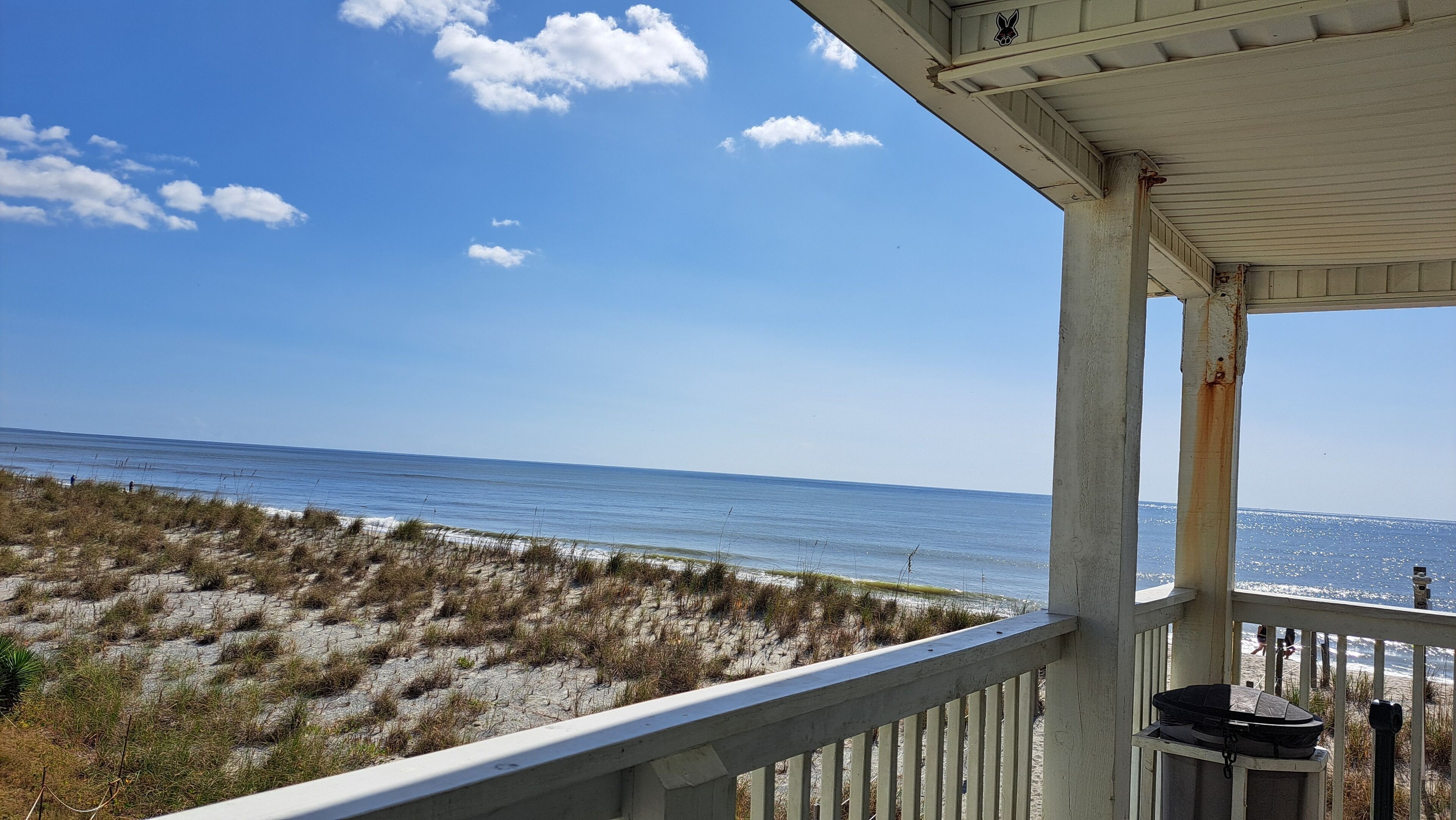 A beach view from a covered porch with a railing, overlooking the ocean and sandy dunes.