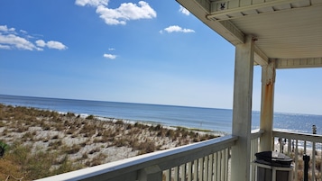 A beach view from a covered porch with a railing, overlooking the ocean and sandy dunes.