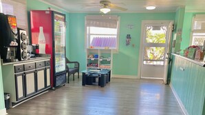 A spacious room with a wooden floor, a refrigerator, a coffee machine, and a window with blinds. - Ocean Crest Motel (Oak Island)