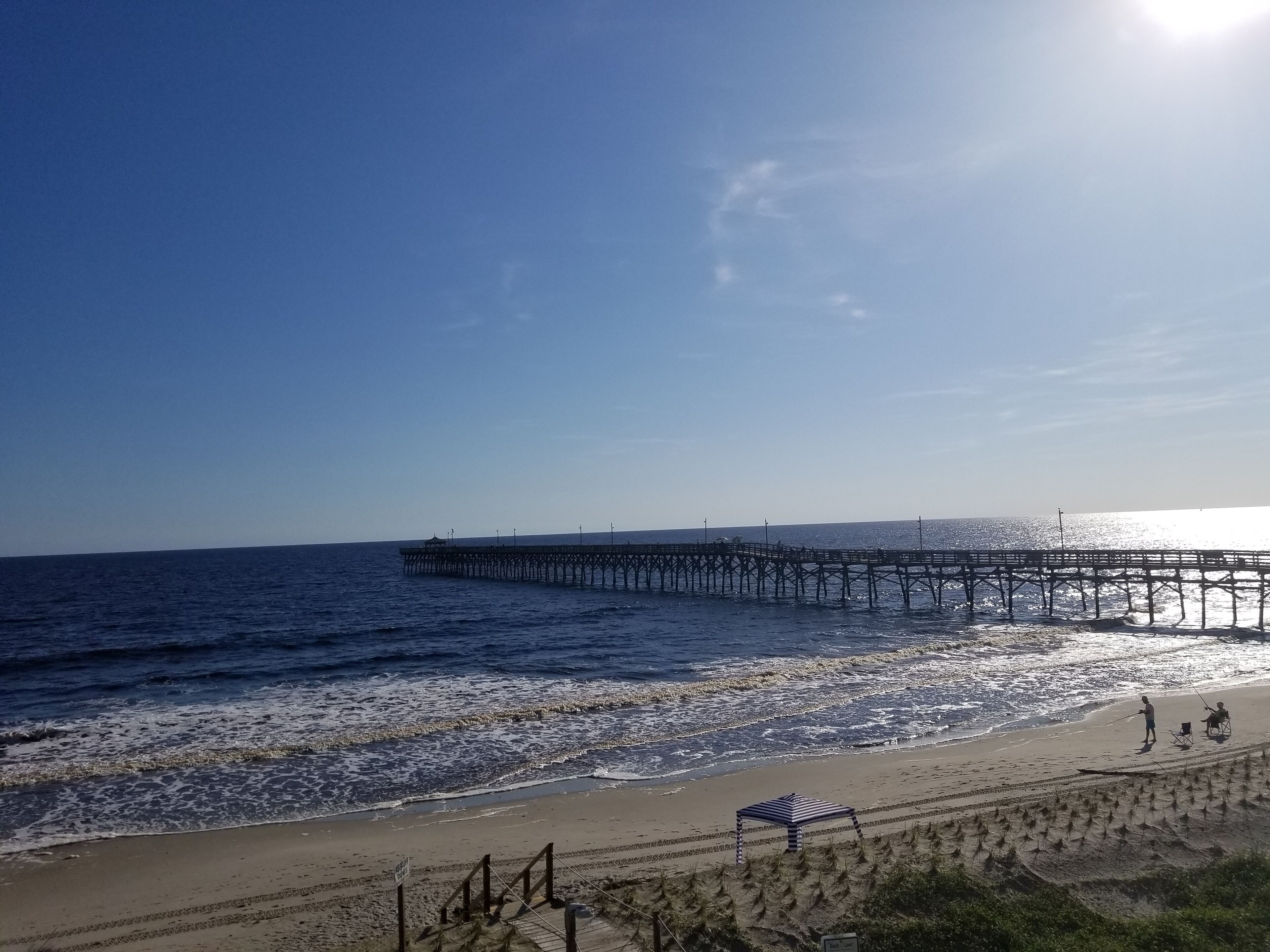 En strand med en träbrygga som sträcker sig ut i havet, en sandstrand och ett randigt strandparasoll.