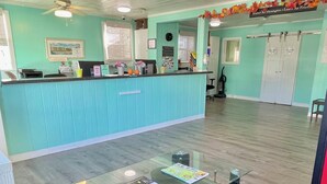 Une pièce avec une table à plateau en verre, des chaises noires, un comptoir d’accueil de couleur turquoise, un ventilateur de plafond et un panneau sur le mur.