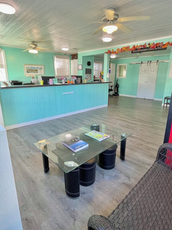 A room with a glass-top table, black chairs, a teal-colored reception desk, a ceiling fan, and a sign on the wall. - Ocean Crest Motel (Oak Island)
