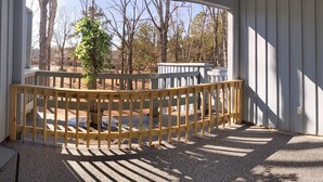 A covered outdoor area with a wooden railing, a small table, and a view of trees and buildings in the background. - Comfy Corner a large comfortable  one room condo with a strict no smoking policy (Fairfield Harbour, New Bern)