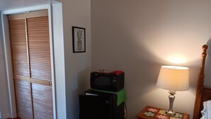 A room with a wooden floor, a closed door, a microwave on a green cloth, a wooden nightstand with a lamp, and a small trash can. - Comfy Corner a large comfortable  one room condo with a strict no smoking policy (Fairfield Harbour, New Bern)