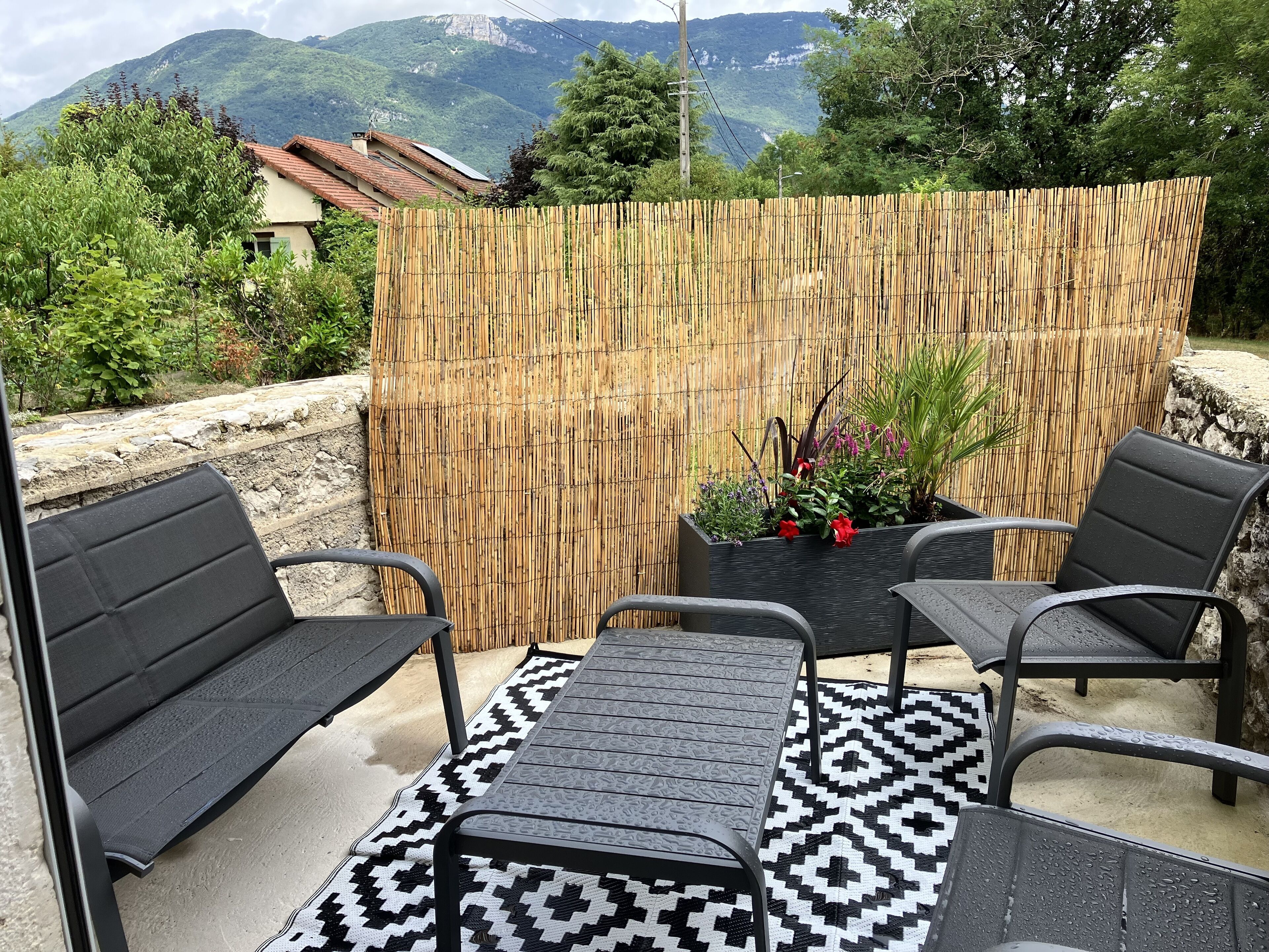 An outdoor patio with two black metal chairs and a table, a patterned rug, and a bamboo fence.