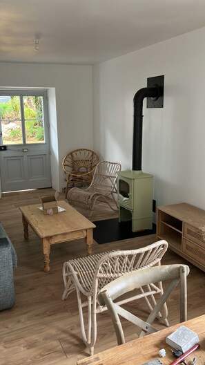 A living room with a wooden floor, a green wood-burning stove, wicker furniture, and a door leading outside.