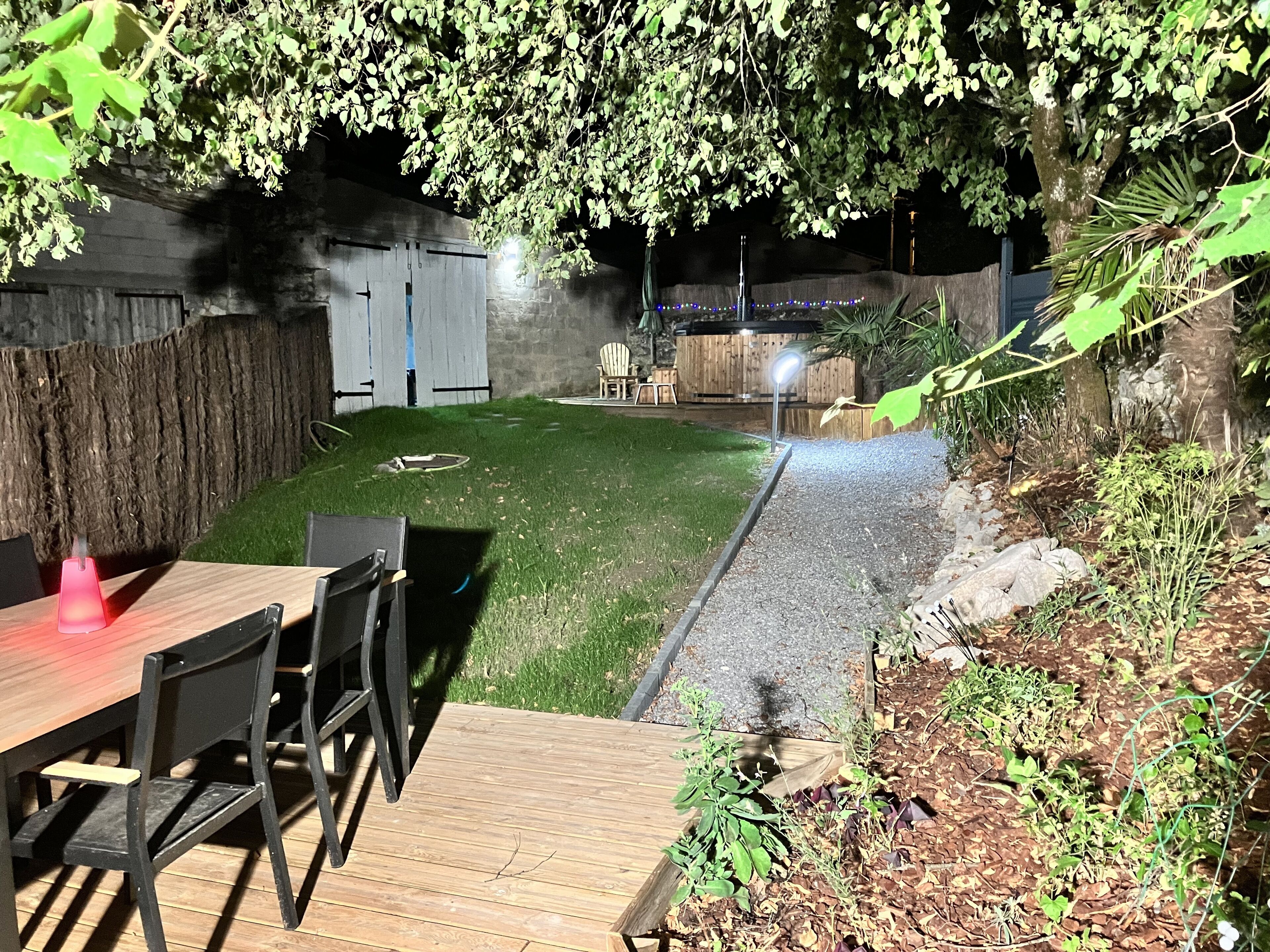 Une terrasse en bois avec une table et des chaises, entourée d’un jardin comprenant une pelouse et un chemin de gravier.