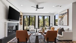 A modern living room with a fireplace, a flat-screen TV, a ceiling fan, and a large window with a view of trees. - Abode at Hollow Mountain, private hot tub, sound system, garden (Park City)