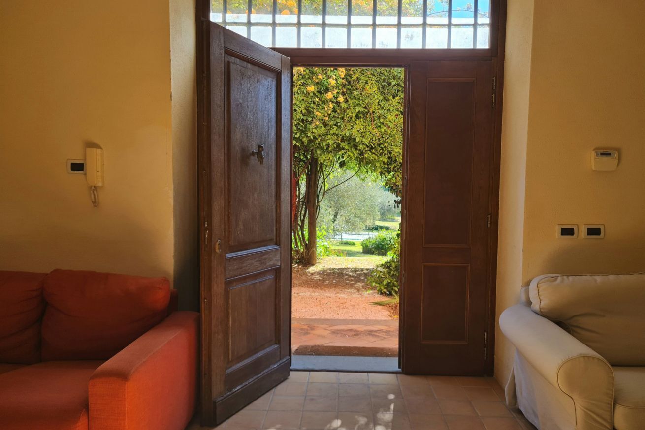 Una stanza con una porta in legno che dà su un giardino, un divano rosso e un divano color beige.