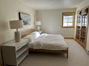 Ett sovrum med en säng, två sängbord, en lampa, ett litet bord, en hylla, ett fönster med gardiner och en dörr med glaspaneler.