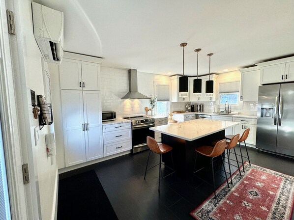 A modern kitchen with white cabinetry, a central island with bar stools, stainless steel appliances, and pendant lighting. - Renovated Gem Near Cantine Field, HITS & Hunter (Saugerties)