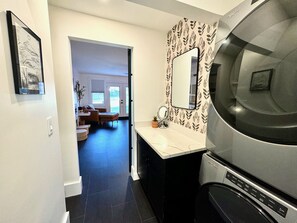 A modern kitchen with a built-in washer and dryer, a large mirror, and a dining area visible through an open door. - Renovated Gem Near Cantine Field, HITS & Hunter (Saugerties)