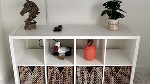 Un mueble blanco para televisor con un televisor de pantalla plana montado encima. El mueble tiene tres estantes, cada uno con una cesta tejida.