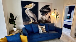 Una sala de estar con un sofá azul, un sillón amarillo, un cuadro grande con pájaros, una planta en maceta y una lámpara de pie.