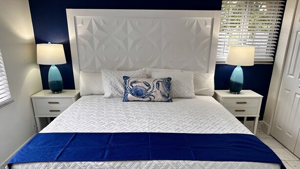 Una cama bien tendida con cabecera blanca y ropa de cama azul, flanqueada por dos mesitas de noche con lámparas, en una habitación con una pared de acento azul oscuro y una ventana con persianas.