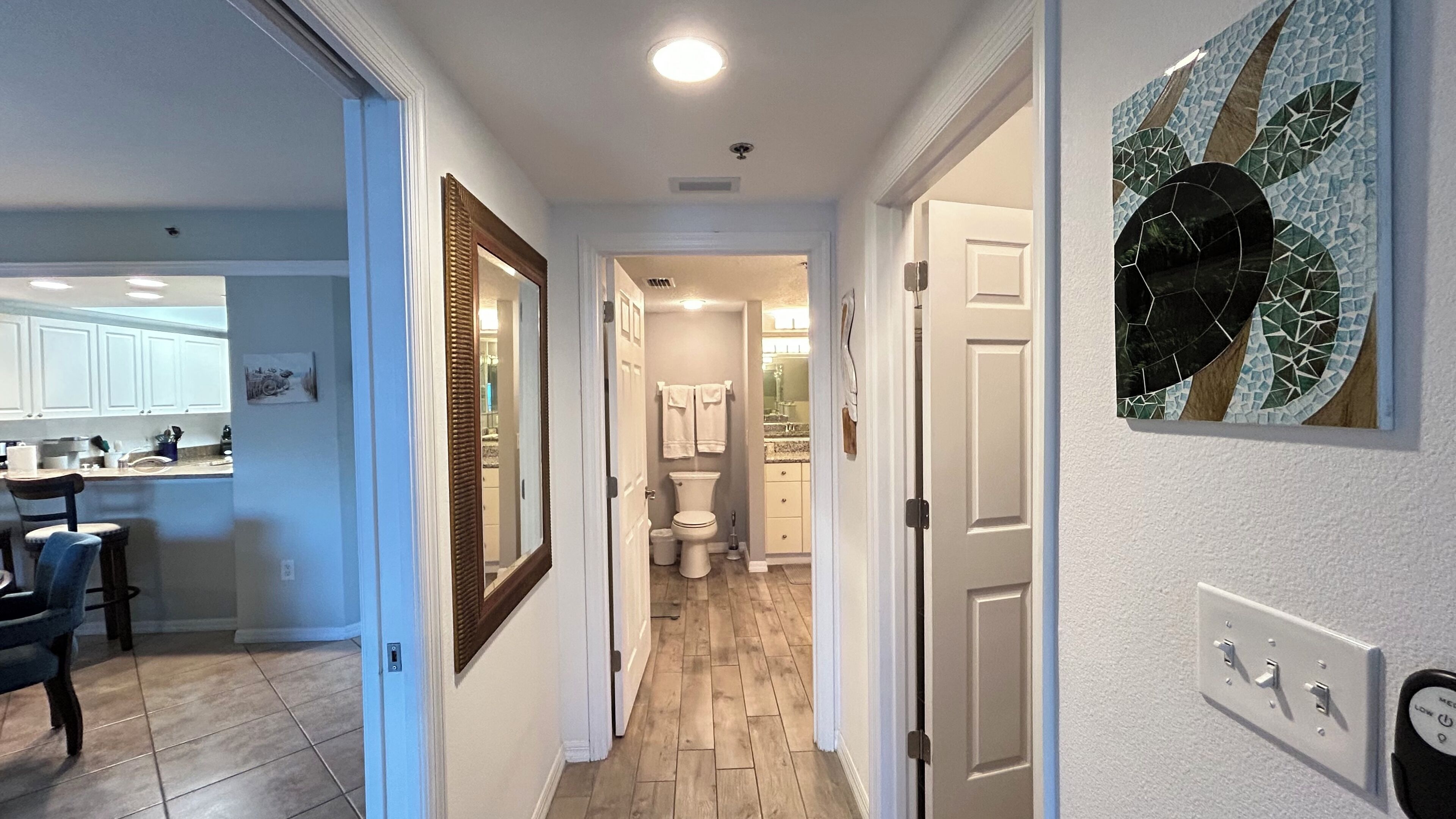 Un couloir mène à une salle de bain équipée de toilettes, d’un lavabo et d’un miroir. Il y a une porte sur la droite et une chaise sur la gauche.