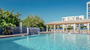A clear blue pool with a pergola and lounge chairs, surrounded by trees and a building with balconies. - Seashell Escape (New Smyrna Beach)