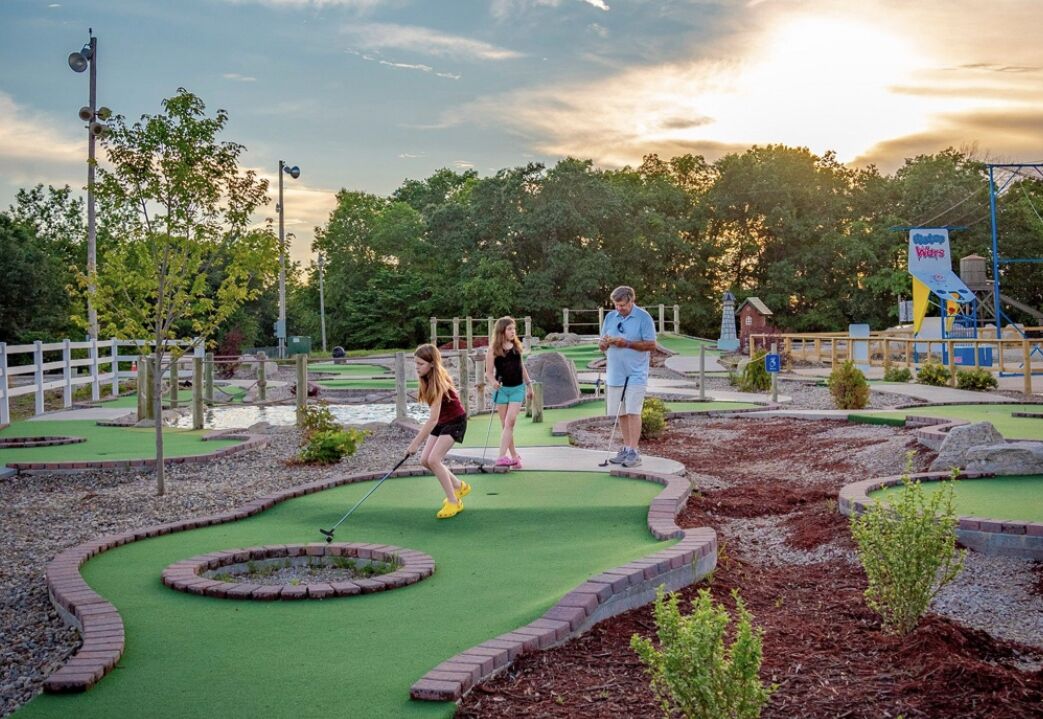 A miniature golf course with a white fence, green grass, and a few trees in the background.