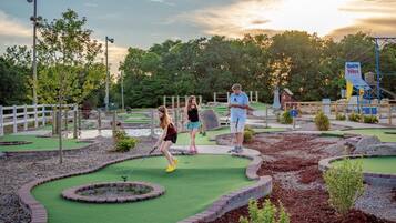 A miniature golf course with a white fence, green grass, and a few trees in the background.