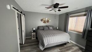 Un dormitorio con una cama, un ventilador de techo, una mesita de noche, un reloj de pared, una ventana con persianas y una puerta con cortina.